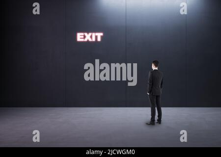 Create your future and best way-out concept businessman back view in empty room looking at glowing neon exit sign on dark wall Stock Photo