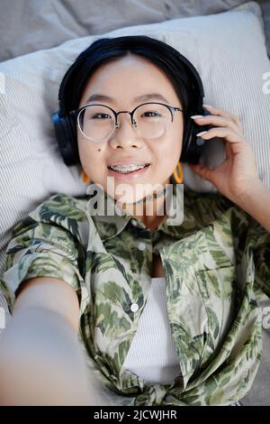 Vertical POV shot of Asian teenage girl listening to music in headphones and smiling to camera while lying on bed and filming video Stock Photo