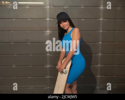 Young Hispanic woman in trendy activewear and cap with skateboard looking at camera while standing near metal wall on weekend day in city Stock Photo