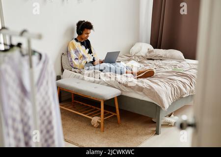 Wide angle view at black teenage boy with laptop sitting on bed in messy room, copy space Stock Photo