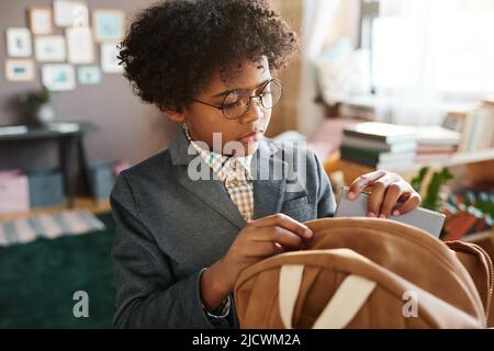 African school boy in eyeglasses and formal suit packing books in his school bag, he preparing for school at home Stock Photo