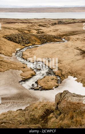 Mountain stream flowing from the snow in the hills and mountains near Hvammstangi in Iceland towards the fjord in the distance Stock Photo