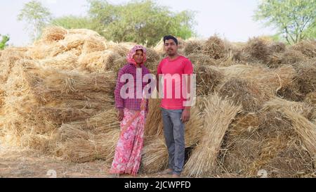 Young Indian farmer standing with his wife in front of wheat crop pile. Stock Photo