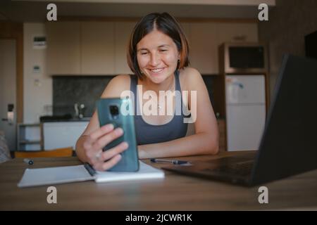 Busy young woman talking on smart phone with clients, coworkers, working remotely on laptop in morning. Female freelancer tutor student having phone conversation call. Stock Photo