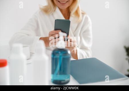 Blonde mature woman sitting in cosmetology center and uses cellphone surfing the net Stock Photo