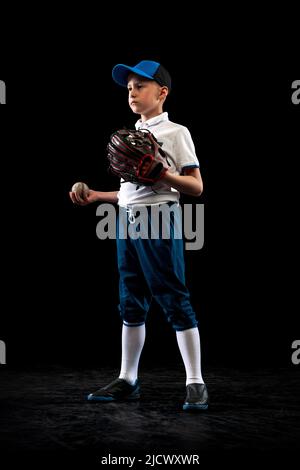 Portrait of kid, beginner baseball player in sports uniform posing with baseball glove isolated on dark background. Concept of sport, achievements Stock Photo
