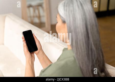 Back view senior gray-haired woman sits on couch and holds gadget with blank display, touching touchscreen with finger, mock-up, app advertising. Woman holding smartphone with an empty screen Stock Photo