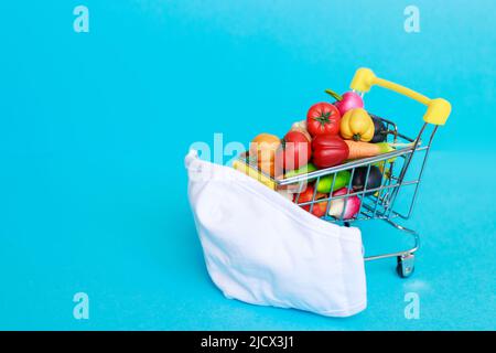 Shopping cart with protective medical mask with fruits and vegetables on a blue background. Safe and online shopping on quarantine concept. Stock Photo