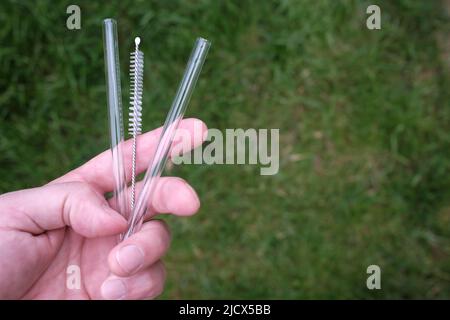 Hands hold reusable glass straws for cocktails and a special cleaning brush, on a background of greenery. Ecological concept.  Stock Photo