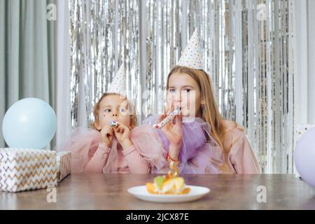 Two sisters of 5 and 11 years old in party hats and elegant dresses are sitting at table and blowing holiday horns, with cake in front of them Stock Photo