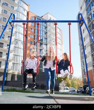 Cheerful siblings sitting on swing, while happy mum carefully pushing behind at playground. Woman swinging cute daughter and son at modern courtyard of city residential high-rise buildings. Stock Photo