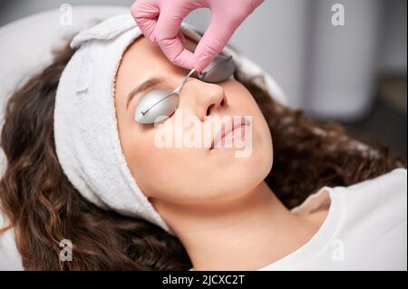 Close up of cosmologist hand putting protective goggles on women eyes before beauty procedure in cosmetology clinic. Young woman wearing headband and safety glasses while resting in beauty salon. Stock Photo