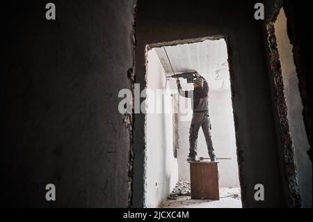 Full length of man builder standing on wooden table and using electric drill while making hole in wall. View through doorway of male worker drilling wall in room under renovation. Stock Photo