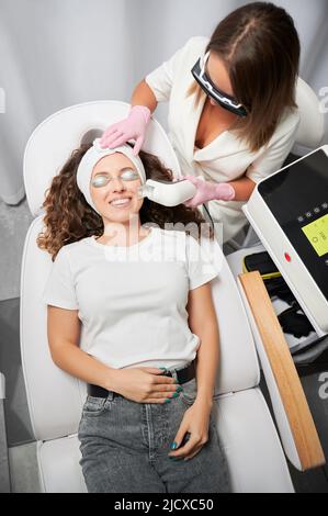 Smiling young woman in protective goggles receiving laser skincare treatment in cosmetology clinic. Female beautician using diode laser scanner device while performing facial skincare procedure. Stock Photo