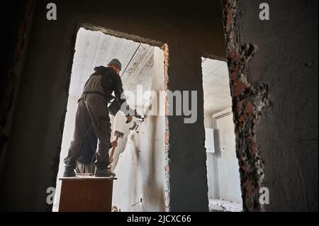 Full length of male builders using hammer drill and sledgehammer while destroying wall in building. View through doorway of two workers breaking wall in apartment under renovation. Stock Photo