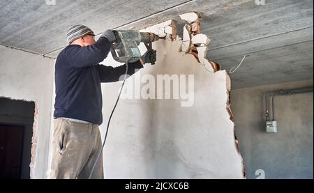Man builder drilling wall with electric hammer drill. Male worker using professional drilling tool while breaking wall in apartment during renovation. Concept of home renovation and reconstruction. Stock Photo