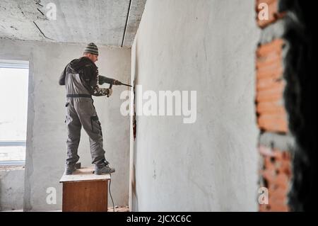 Full length of man builder standing on wooden desk and drilling wall with special construction tool. Male worker using electric drill machine while breaking the wall in apartment during renovation. Stock Photo