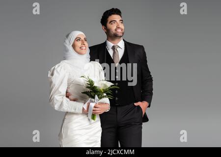 groom in suit posing near muslim happy bride with wedding bouquet on grey Stock Photo
