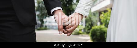 cropped view of bride in wedding ring holding hands with groom outdoors, banner Stock Photo