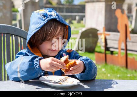 Little boy, 4 years old, enjoying a cake at an outdoor cafe on a chilly day at Portchester Castle, Hampshire, UK.  MODEL RELEASED Stock Photo