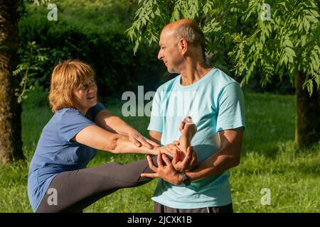 Mature couple in the green park stretching legs together while exescising to improve their health in elder ages Stock Photo