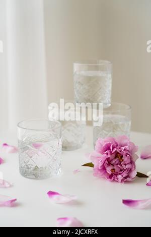 faceted glasses stacked near pink peony and petals on white tabletop and grey background Stock Photo