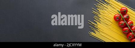 Top view of cherry tomatoes on branch on uncooked pasta on black background, banner Stock Photo