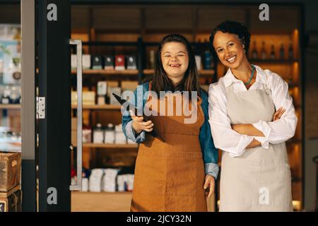 Two successful store employees smiling at the camera while standing at the entrance of their grocery store. Happy woman with Down syndrome working in Stock Photo