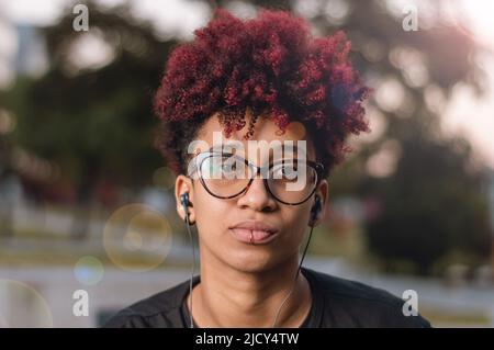 close up portrait outdoors beautiful young latin brunette woman with dyed red afro, glasses and headphones, serious looking at the camera Stock Photo