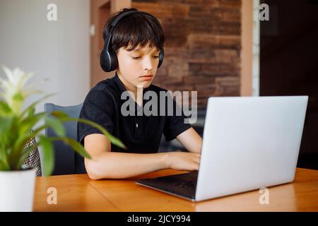 Portrait of teenager boy wear black T-shirt, headphones, sitting at desk, typing on laptop, surfing internet, chatting. Stock Photo