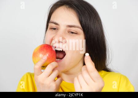 A woman bites a red apple while holding a multivitamin pill in her hand Stock Photo