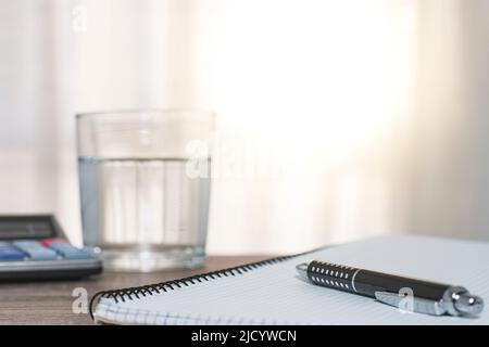 On a wooden table there is a calculator, a glass of water and a notebook with checkered sheets on which there is a pen. Stock Photo
