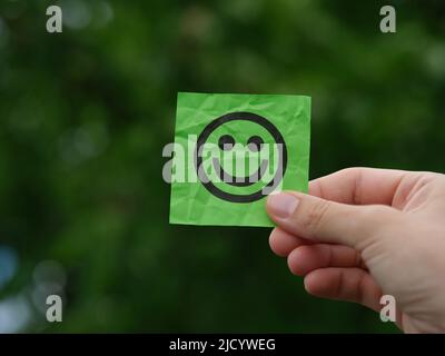 A woman holding a green sticky note with a happy face on it in her hand against a background of foliage. Close up. Stock Photo