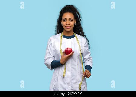 African American Woman Doctor Nutritionist wears White Coat. Good Looking Dietitian in Medical Uniform with Red Apple and Measuring Tape on Blue Studi Stock Photo