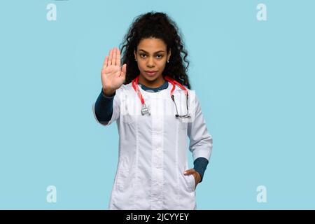 Serious African American Female Physician Medical Advisor Shows  Stop Palm Sign on Blue Studio Background. Smiling Woman Doctor GP Wears Hospital Unif Stock Photo