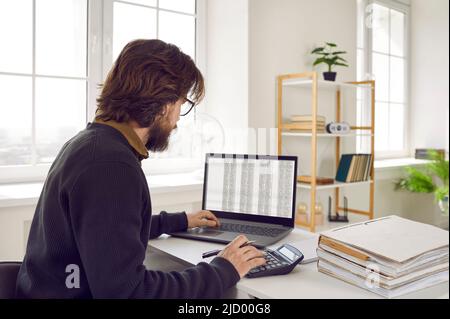Male accountant sitting in office, working with spreadsheets on laptop and using calculator Stock Photo