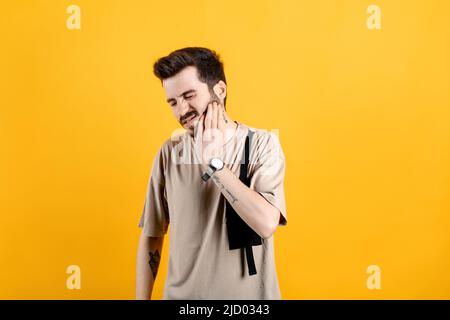 Young man wearing beige t-shirt posing isolated over yellow background feeling pain, holding his cheek with hand, suffering from bad toothache. Painfu Stock Photo