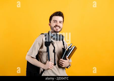 Portrait of man wearing beige tee posing isolated over yellow background student with books and backpack and looking at the camera. High school univer Stock Photo
