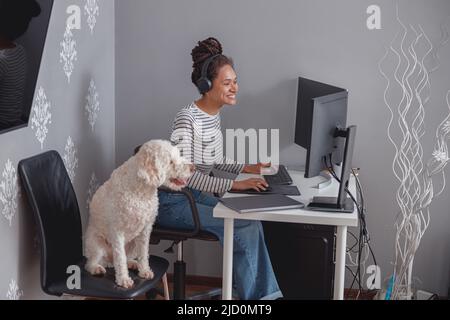 Young happy mixed-race woman programmer in headphones working on computer at home Stock Photo