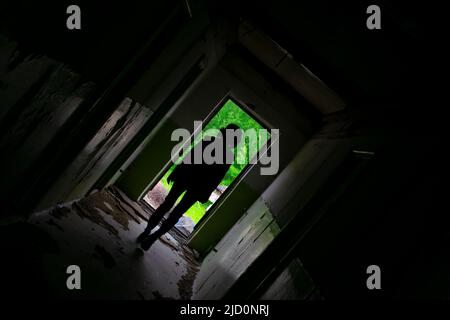 a view of a dark sloping corridor of an abandoned building with a silhouette of a girl walking to the exit, in which the bright green foliage of trees Stock Photo