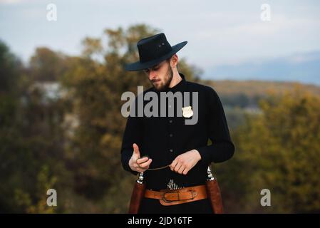 Sheriff or cowboy in black suit. Man with vintage pistol revolver and marshal ammunition. American western Sheriff. Wild west with cowboy. Stock Photo