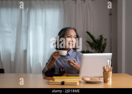 Happy joyful mature laptop user asian woman chatting online, typing, making video call over cup of coffee, tea, relaxing on couch. Senior freelance Stock Photo