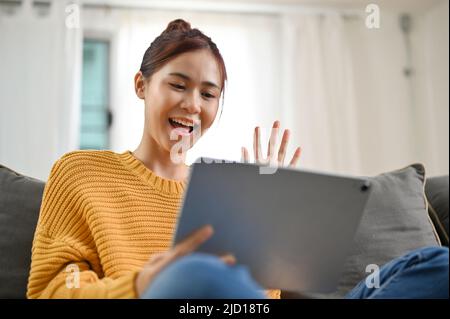 Cheerful young asian woman video calling with her friends, say hi or hello through the web cam, live streaming on social media via tablet. Stock Photo