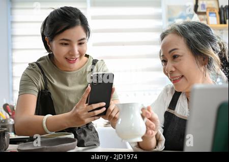 An attractive asian young woman using smartphone, taking a picture of her mom's ceramic jar in the workshop. Stock Photo