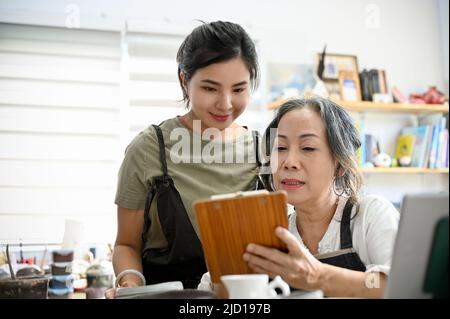 Professional asian aged woman coaching and showing the moulding clay instruction on the paper to young female student in the workshop. Stock Photo