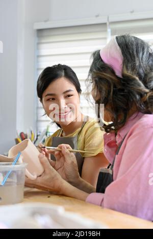 Attractive charming young asian woman talking and painting a ceramic cup with an aged woman in the ceramic pottery workshop. Stock Photo