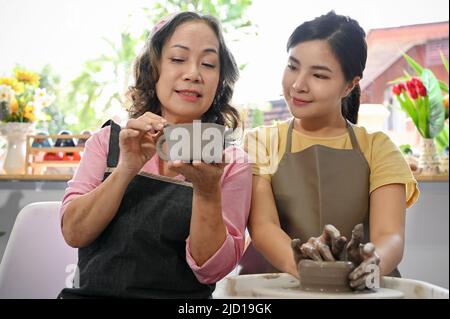 A professional potter teaching and coaching her young female student making a pottery in the workshop. Charming asian woman moulding clay on potter's Stock Photo