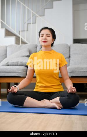 Relaxed calm young Asian woman practicing yoga at home, practicing a lotus pose and meditating on the yoga mat. Stock Photo