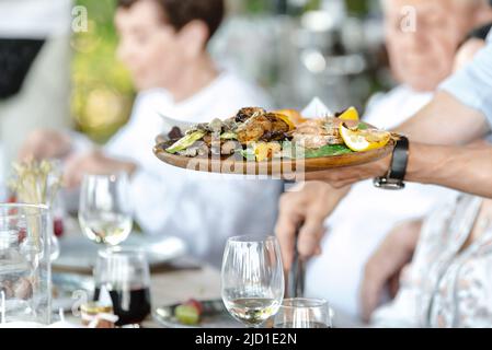 The waiter serves restaurant guests. The waiter holds a tray with food shrimp and vegetables Stock Photo