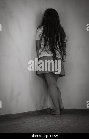 Black and white portrait of little frightened girl with long hair standing punished in corner in empty room. Vertical. Stock Photo